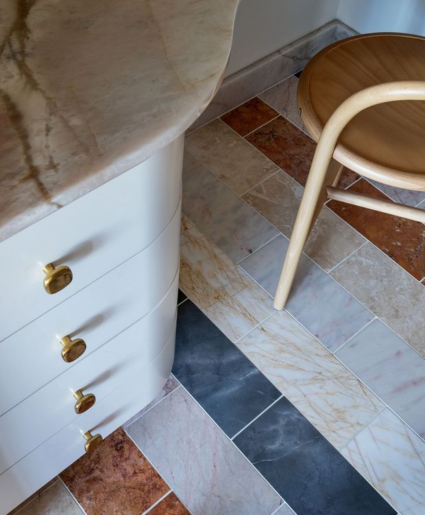 Close-up interior detail showing a curved white built-in vanity or cabinet with rounded drawers and brass knobs, paired with a pale stone countertop. The floor is composed of varied rectangular natural-stone or stone-look tiles in cream, taupe, terracotta, pink, and dark charcoal tones. A light timber-framed curved chair is partially visible at right. The composition highlights sculptural curved forms, mixed stone surfaces, and warm brass hardware.