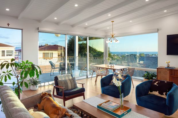 Bright coastal living room with large corner glazing overlooking the ocean and neighbouring homes. The room features a light timber floor, white painted walls and exposed white ceiling beams, a neutral sofa, deep blue lounge chairs, a timber coffee table, indoor plants, and a mid-century-inspired pendant light. A balcony or terrace is visible beyond the glazing.