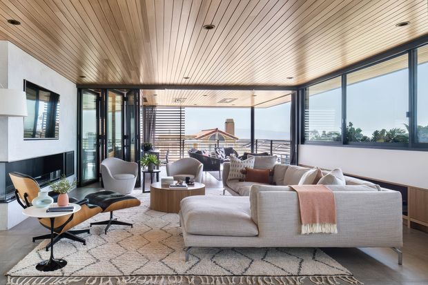 A bright, contemporary living room with expansive black-framed glazing opening onto a balcony or terrace. The room features a large pale sectional sofa, sculptural lounge chairs, a round coffee table, patterned area rug, timber slatted ceiling, light walls, and dark built-in joinery. A coastal outlook and outdoor seating are visible beyond the glazing.