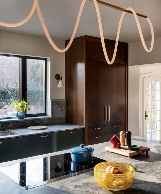 A refined kitchen with dark green lower cabinetry, a tall dark timber pantry or storage wall, and a pale veined stone benchtop. A large black-framed window overlooks greenery, while a sculptural looping pendant light spans the ceiling. The island includes a black induction cooktop and is styled with a blue Dutch oven, fruit bowl, and serving tray. A glazed exterior door is visible beyond the kitchen.