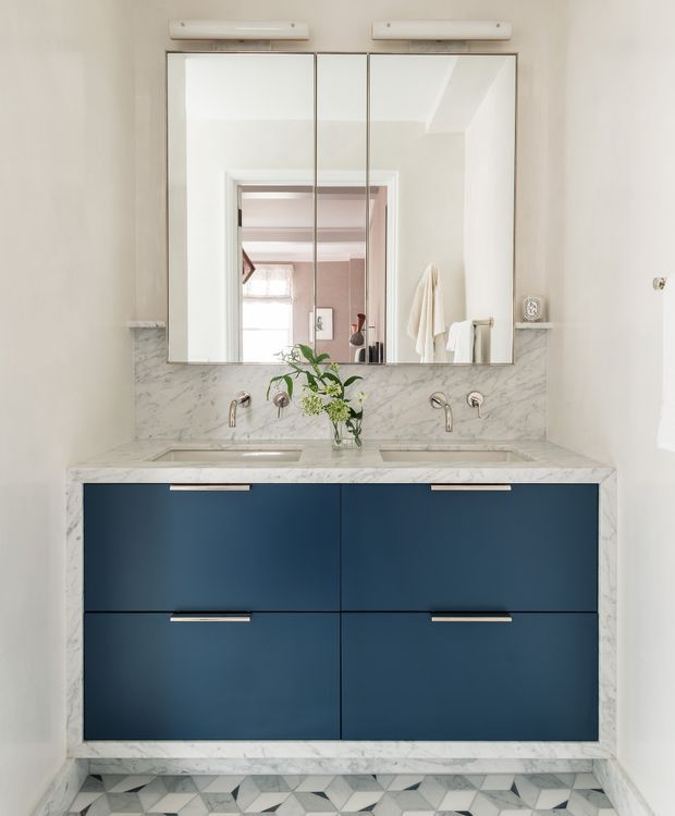 A refined bathroom vanity with a double sink, deep navy-blue floating cabinetry, white marble benchtop and backsplash, wall-mounted chrome fixtures, and a large mirrored cabinet above. Geometric patterned floor tiles and warm illuminated mirror lighting add visual interest. The mirror reflects an adjacent pink-toned room and towels, while the main space remains bright and clean.