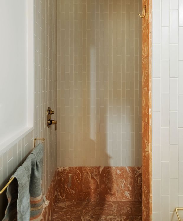 A compact bathroom shower enclosure viewed straight on. The shower walls are clad in narrow vertical beige tiles, with a lower band and side trim of richly veined reddish-brown stone. Brass shower controls, hooks, and towel rail add warm metallic accents, while a pale towel hangs at the left.