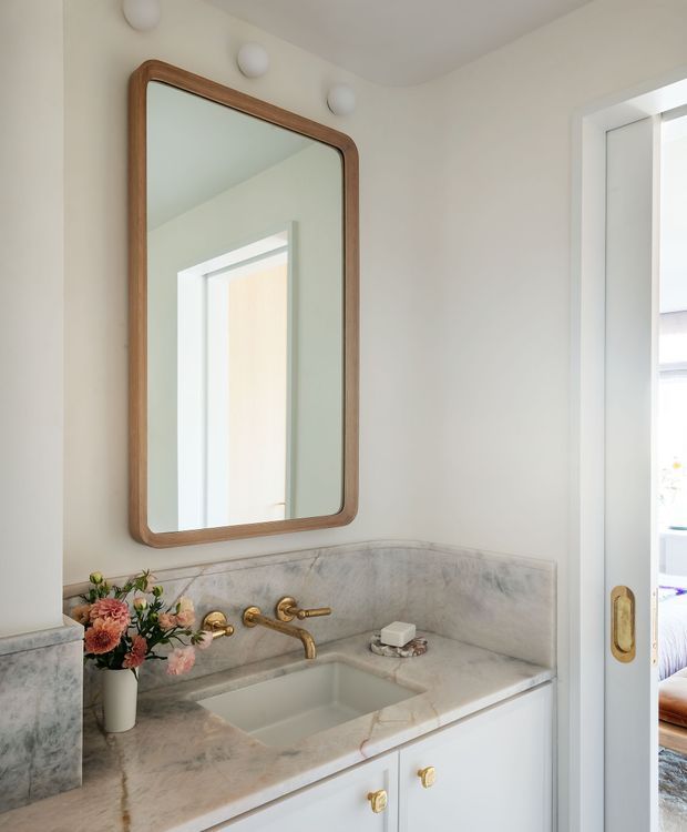 Bright, refined bathroom vanity with a white marble countertop and integrated backsplash, undermount rectangular basin, brushed brass wall-mounted tapware, and white cabinetry with brass pulls. A large rounded-corner timber-framed mirror sits above the vanity, accompanied by globe wall lights. A partially open pocket door reveals an adjacent bedroom or living space. The palette is predominantly white and pale grey with warm timber and brass accents.