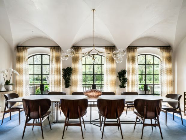Bright, elegant dining room with a long rectangular dining table surrounded by sculptural dark wood dining chairs. Three tall arched windows with black muntins and sheer patterned curtains provide abundant natural light and views of greenery. The room features a white vaulted, barrel-like ceiling with repeated arches, a contemporary multi-globe pendant light, pale flooring, indoor plants, and refined neutral furnishings.