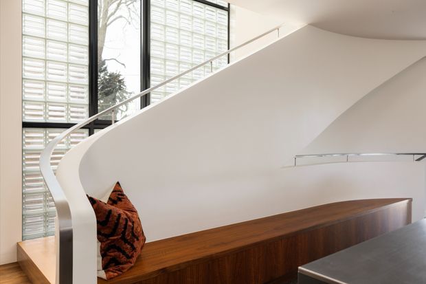 Bright contemporary interior focused on a sculptural curved white staircase with a slim glass or clear balustrade. The staircase rises beside tall glazing and a translucent glass-block wall, with a long dark timber built-in bench or cabinet beneath it. A small patterned cushion sits at one end, and a dark stone or composite benchtop is visible in the foreground. The space is clean-lined, open, and strongly daylit.