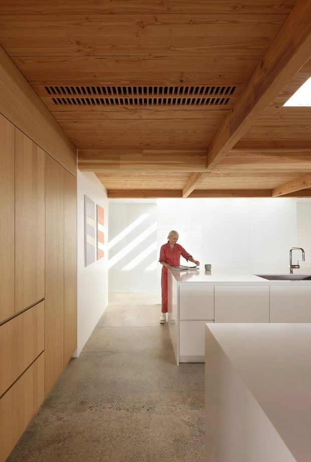 Bright contemporary kitchen with a warm timber-lined ceiling featuring exposed beams and a linear ventilation grille. Full-height timber cabinetry lines the left wall, while a white island and white cabinetry with a pale benchtop occupy the right side. A stainless-steel tap and integrated sink are visible, with polished concrete flooring and strong sunlight entering from the rear.