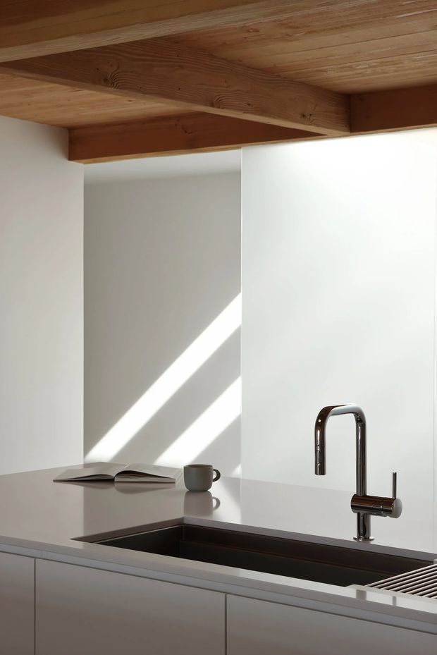 A minimalist kitchen interior focused on a pale stone or engineered-stone benchtop with an inset dark sink and polished chrome gooseneck tap. White walls and handleless light cabinetry create a clean, restrained composition, while exposed warm timber ceiling boards and a substantial timber beam add natural contrast. Strong diagonal sunlight falls across the white wall, with an open book and cup resting on the benchtop.