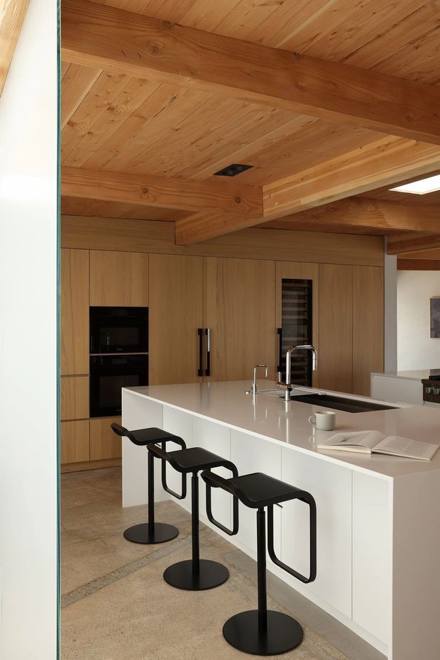 Contemporary kitchen with a large white waterfall island, integrated sink and appliances, light timber cabinetry, and three black minimalist bar stools. The room features an exposed timber-lined ceiling with substantial timber beams, recessed lighting, and a neutral stone or concrete-look floor.