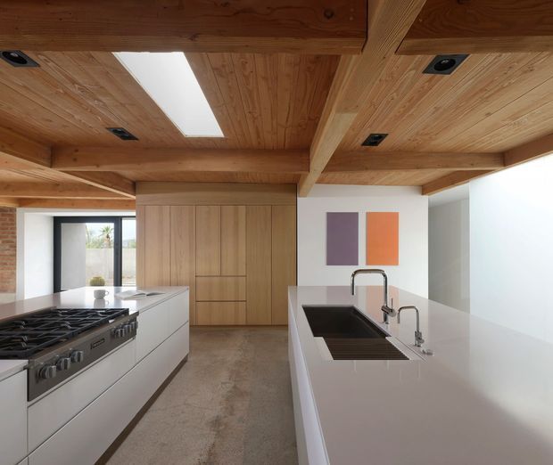 Contemporary kitchen with a large central island, integrated dark sink, twin faucets, white cabinetry and a professional stainless-steel range. Warm timber lining covers the ceiling and exposed beams, with a long skylight providing daylight. Full-height timber pantry or storage joinery sits against a white wall, while exposed brick is visible at the left edge and two colourful abstract panels add accents.