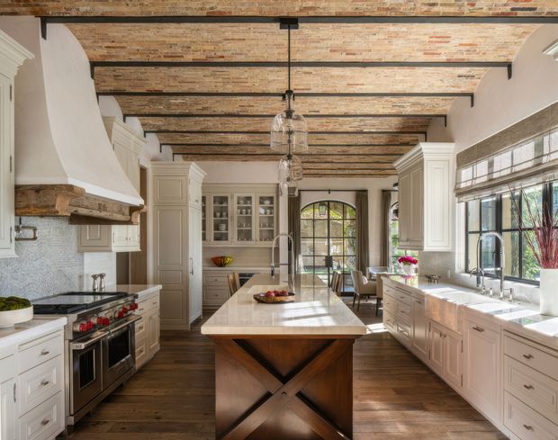 A spacious, refined kitchen with a large central island, extensive cream-painted cabinetry, dark timber flooring, a professional stainless-steel range, and a broad bank of windows over the sink. The room features a distinctive exposed brick ceiling with dark structural beams, an arched glazed door, pendant lights, and a substantial timber island base with crossed detailing.