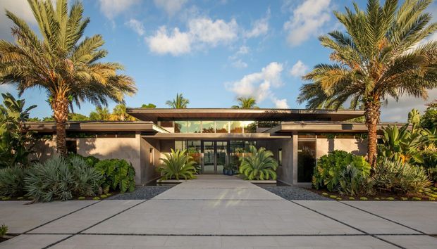 Front exterior view of a low, single-storey modern residence with a broad flat roof, deep horizontal overhangs, and a central glazed entry. The facade combines warm beige rendered walls, dark structural and window frames, and extensive floor-to-ceiling glazing. A wide concrete-paved approach is bordered by gravel beds and lush tropical planting, including prominent palm trees and agave-like plants. The composition is symmetrical and strongly horizontal, with a bright blue sky and warm sunlight.