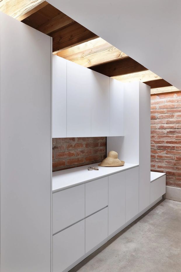 A minimalist built-in storage and wardrobe installation in a narrow interior space. Floor-to-ceiling matte white cabinetry with integrated drawers and cupboards surrounds a white bench or counter, set against exposed red brick walls. Exposed timber ceiling beams and a polished grey concrete floor add warmth and industrial character. A straw hat and eyeglasses rest on the counter.