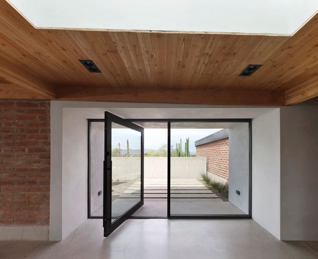 A minimalist interior opening onto a small paved courtyard through black-framed floor-to-ceiling glazing and an open hinged glass door. The room has a smooth pale concrete floor, white plastered walls, an exposed timber-lined ceiling with recessed black spotlights, and exposed red brick side walls. The courtyard continues the restrained material palette with concrete paving, a rendered boundary wall, brickwork, and sparse planting.