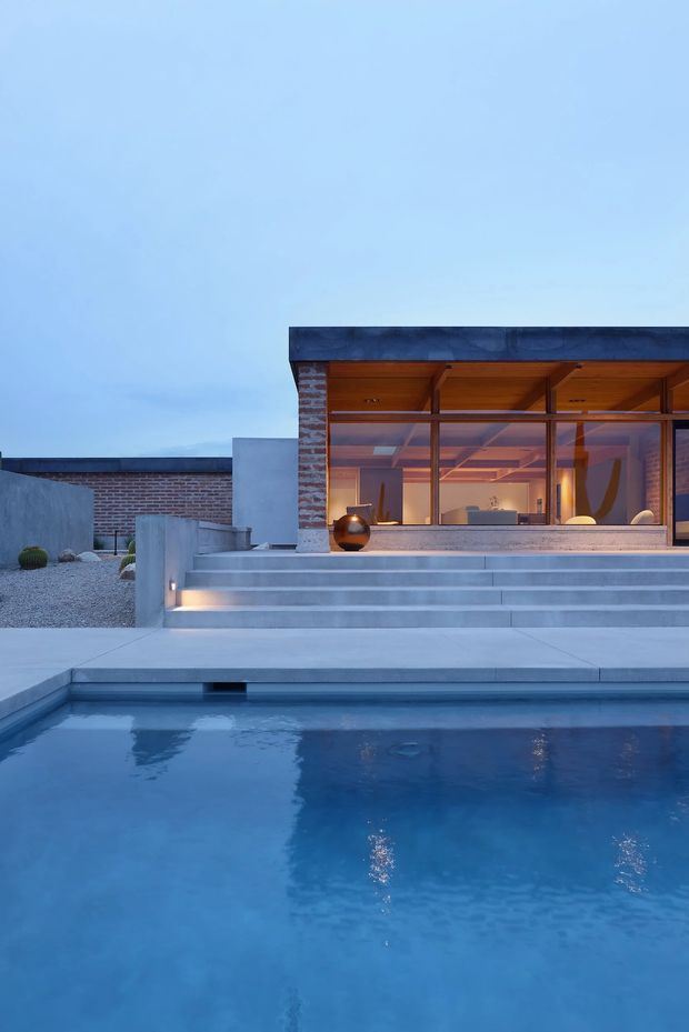 A dusk exterior view of a contemporary residential pool courtyard. Broad pale concrete steps rise from the swimming pool to a glazed pavilion with warm timber soffits and exposed structural beams. Brick walls, dark flat roof edges, frameless-looking glazing, gravel landscaping, and minimalist concrete forms create a clean modern composition.