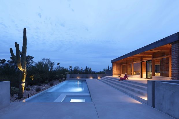 Twilight exterior view of a modern single-storey residence with a long flat roof, warm timber soffit and structural framing, brick walls, expansive glazing, and broad concrete steps leading to a rectangular illuminated swimming pool. The property is set in a desert landscape with prominent cacti and sparse planting.