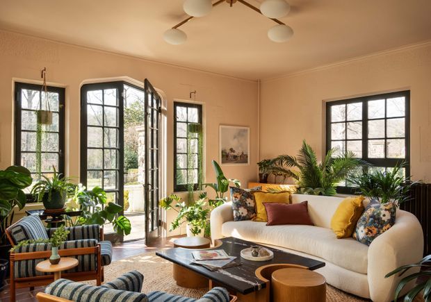 Warm, sunlit living room with tall black-framed multi-pane windows and glazed French doors, cream walls, a large curved cream sofa, patterned and striped lounge chairs, timber nesting stools, and abundant indoor plants. A sculptural multi-globe ceiling light and colourful cushions add a refined yet relaxed character.