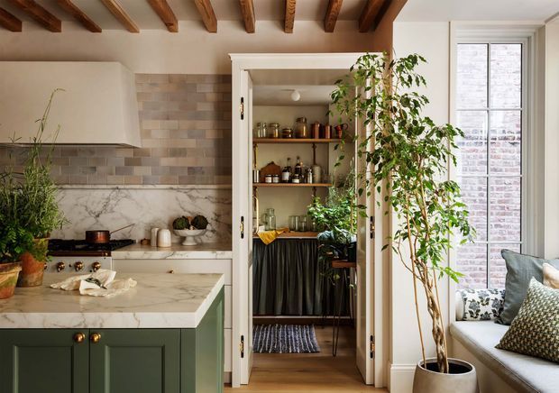 Warm, richly detailed kitchen interior with dark green Shaker-style cabinetry, a pale veined stone island and benchtop, muted rectangular tile splashback, and exposed timber ceiling beams. A tall open pantry or bar storage cupboard sits centrally, with shelves of glassware and bottles, while a bright window seat and large potted tree occupy the right side. The room combines traditional architectural detailing with an inviting, refined contemporary fit-out.