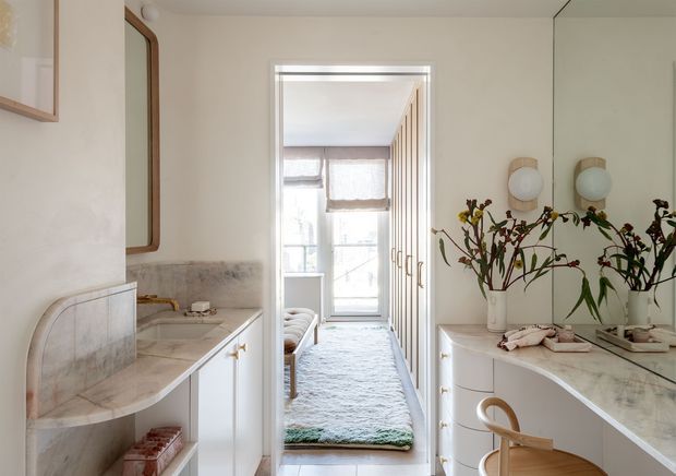 A refined bathroom vanity area viewed toward an adjoining bedroom or dressing space. The bathroom features white built-in cabinetry, a pale marble or stone countertop and backsplash, curved vanity edges, brass hardware, a large wall mirror, and twin round wall sconces. A second vanity is visible in reflection, while the doorway leads to a softly furnished bedroom with a textured rug and tall windows. The overall scheme is light, elegant, and softly traditional.