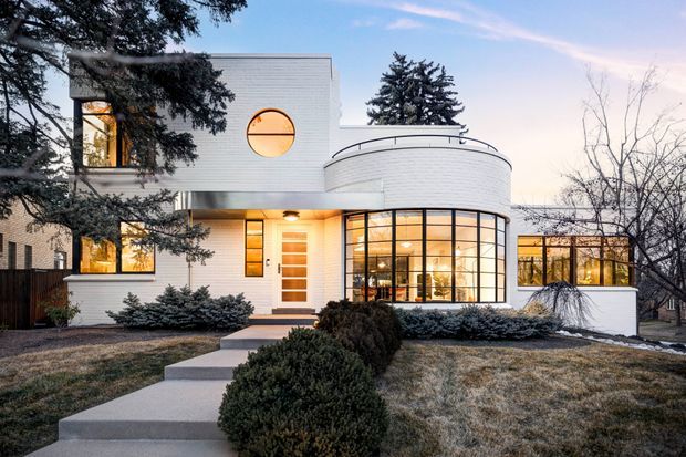 Front exterior view of a striking modern white-brick residence at dusk. The home features a rounded projecting volume with extensive curved black-framed glazing, a circular porthole window, rectilinear upper forms, a warm illuminated entrance, and a stepped concrete path through a landscaped front garden.