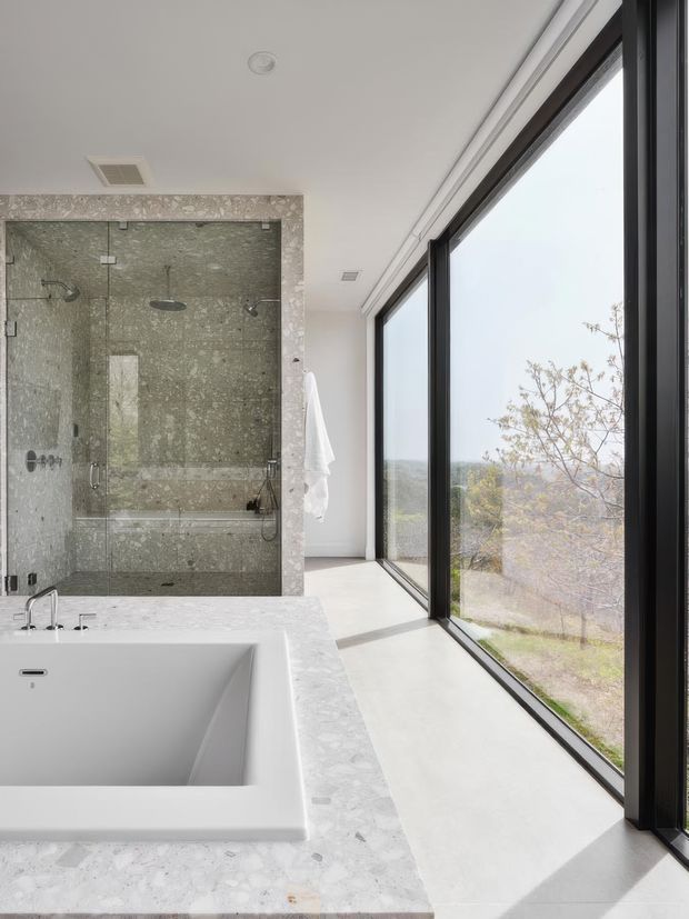 Bright contemporary bathroom featuring a freestanding rectangular bathtub set into a pale stone-clad surround, a large glass-enclosed walk-in shower with matching grey-green stone walls, and expansive black-framed floor-to-ceiling windows overlooking a leafy landscape. The space has a clean, refined aesthetic with abundant natural light and a strong indoor-outdoor connection.