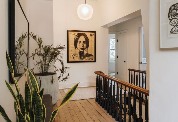 A bright, character-filled upper-floor landing or hallway with pale walls, original timber floorboards, a dark stained timber and turned-baluster stair balustrade, and a large wall mirror reflecting tall indoor plants. Framed artwork and a white globe pendant add decorative interest, while a doorway leads to another room in the background.