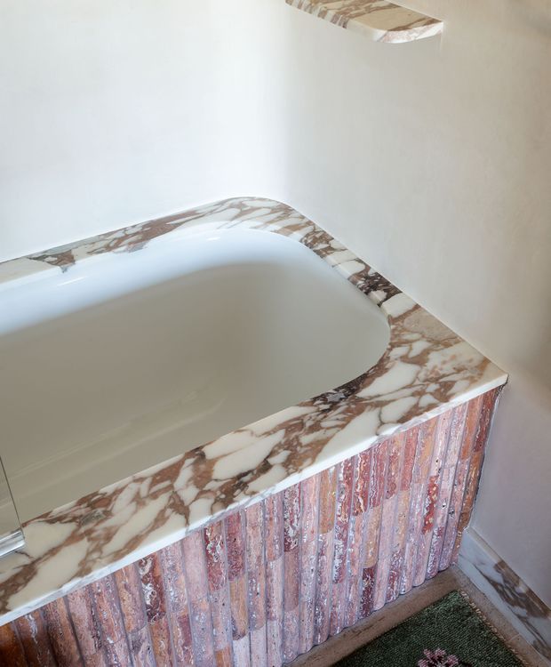 Close-up view of a sculptural built-in bathtub in a bathroom. The tub has a smooth white interior, a rounded rectangular form, and a prominent veined pink-and-white marble rim and surround. The bath front is clad in vertically arranged textured pink-toned tiles or slabs, with matching marble visible at the floor and wall junctions. The overall design is highly decorative and luxurious.