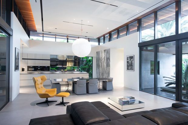 A bright, open-plan contemporary interior combining a minimalist kitchen, dining area and living room. The space features a large white island, integrated white cabinetry, dark framed floor-to-ceiling glazing, high clerestory windows, a polished light floor, a sculptural spherical pendant, and a low dark sectional sofa. Tropical greenery is visible outside through the glazing.