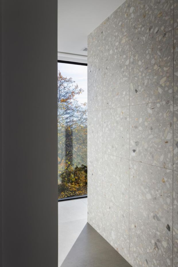 Minimalist interior view featuring a full-height terrazzo or aggregate stone wall with pale grey and beige tones, smooth concrete-like flooring, dark charcoal wall or door edge, and a large window opening to leafy trees outside. The image emphasizes architectural materiality, clean geometric lines, and natural light.