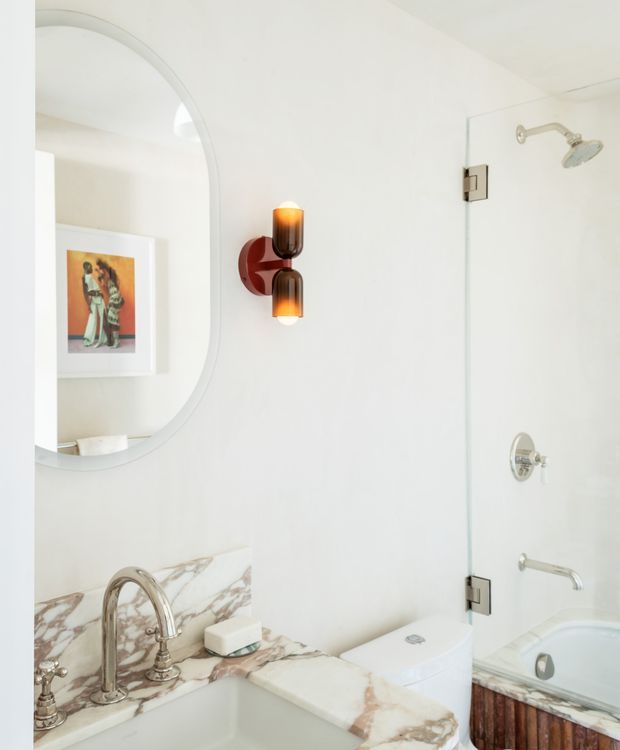 Bright, refined bathroom with a white-painted interior, a large rounded-edge mirror, marble vanity countertop and splashback with prominent brown veining, polished chrome tapware, a wall-mounted amber glass light, and a glass-enclosed shower beside a freestanding-style bathtub. A framed artwork is reflected in the mirror, and timber cladding is visible on the bath exterior.