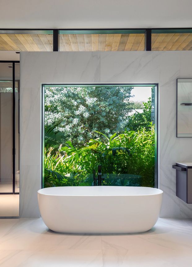 A bright, contemporary bathroom featuring a freestanding white bathtub positioned in front of a large window overlooking dense tropical greenery. The space has pale marble-effect wall and floor surfaces, dark-framed glazing, a floating vanity at right, and a distinctive timber-lined ceiling with dark structural beams.