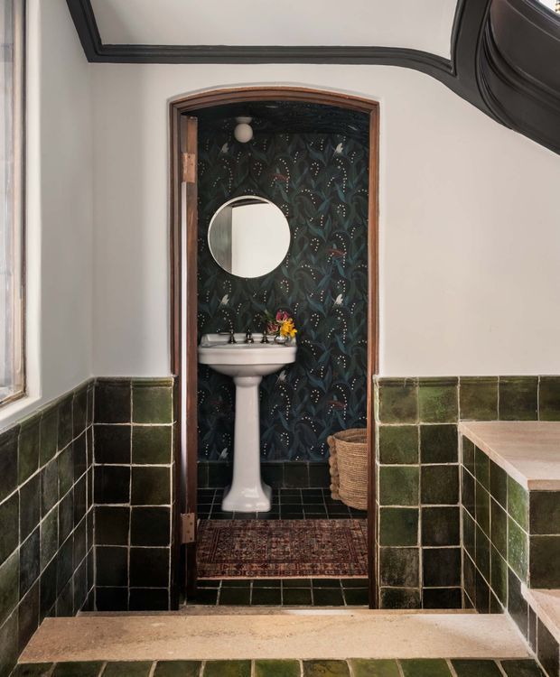 A compact, richly detailed bathroom or powder room viewed through an open doorway. It features a pedestal basin with dark traditional tapware, a round wall mirror, dark patterned feature wallpaper, glossy deep-green wall tiling, a tiled floor, timber-trimmed arched doorway, and a small patterned runner. A curved dark ceiling detail and pale stone or tiled steps frame the foreground.