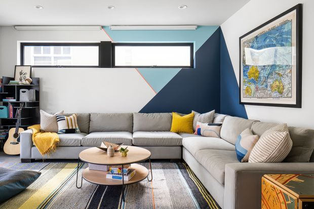 A bright contemporary living room featuring a large L-shaped grey sectional sofa, round two-tier coffee table, patterned area rug, colourful cushions and throws, and a geometric feature wall in white, pale blue, and navy. Narrow horizontal windows provide natural light, while framed artwork and open shelving add personality.