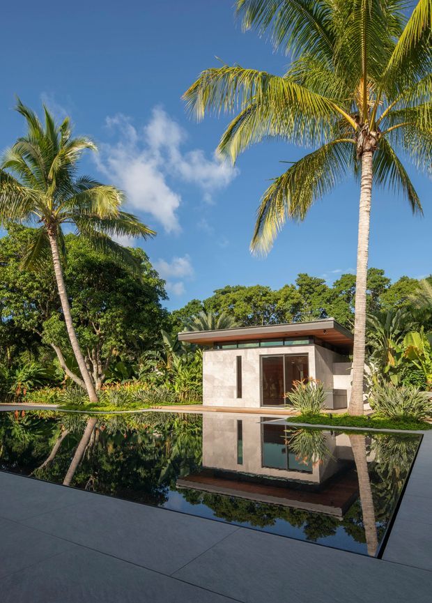 A compact modern pavilion sits beside a highly reflective dark-tiled pool, surrounded by lush tropical planting and tall palm trees. The pavilion has pale stone or concrete walls, a broad shallow roof overhang, timber-toned soffit detailing, and large glazed openings. The scene is photographed in bright sunlight with strong reflections in the still water, creating a serene resort-like outdoor setting.