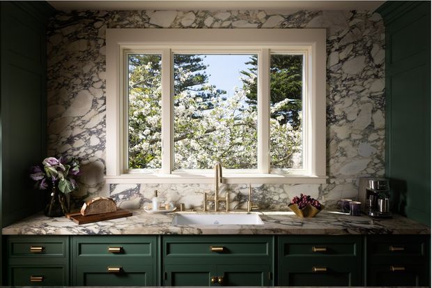 A richly detailed traditional kitchen vignette centred on a three-part timber-framed window overlooking flowering trees. Deep forest-green shaker cabinetry lines the lower half and side walls, while dramatic white-and-grey veined marble wraps the backsplash and benchtop. Brass hardware and a brass-toned gooseneck tap add warmth, with a central undermount sink, coffee appliances, and restrained countertop styling.