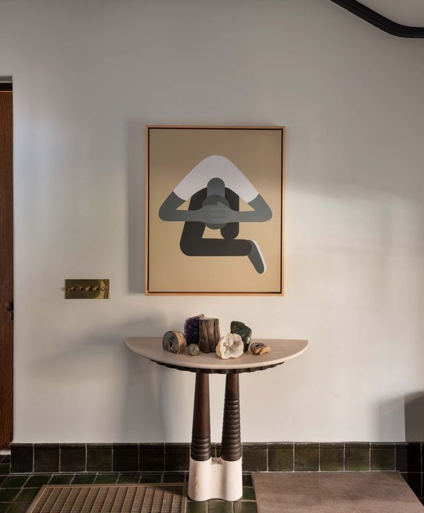 A styled entryway vignette with a narrow sculptural console table against a warm off-white wall. The table has a curved light surface and dark turned supports, displaying small natural sculptural objects. Above it hangs framed abstract artwork, with a dark tiled skirting and timber doorway visible at the side. The space has warm, moody, earthy character with strong decorative styling.