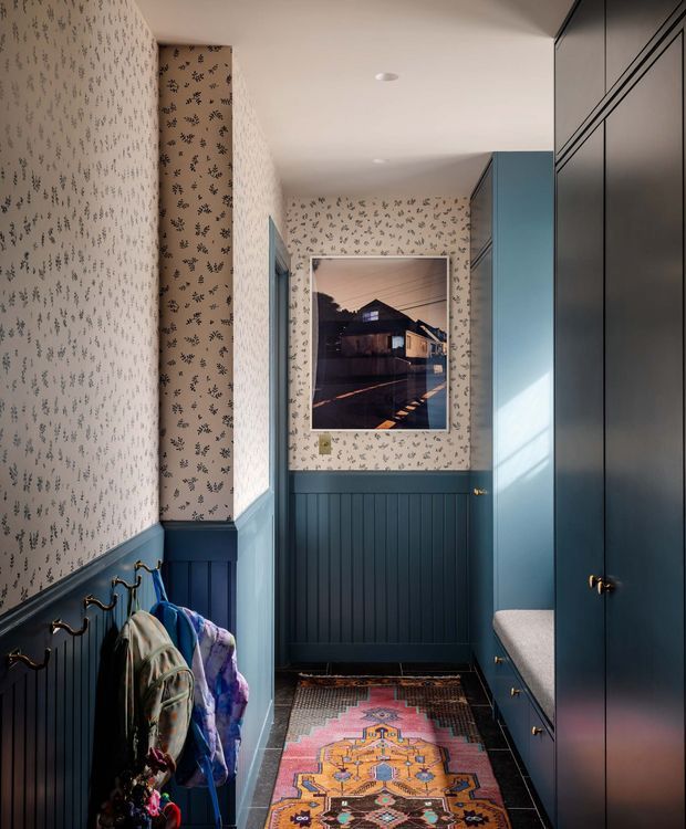 A narrow, richly decorated hallway or entryway with dark blue-green vertical wall panelling, patterned botanical wallpaper, integrated floor-to-ceiling cabinetry, hooks for coats, and a colourful runner rug. A framed architectural photograph is mounted at the far end, while sunlight falls across the cabinetry and a small upholstered bench.