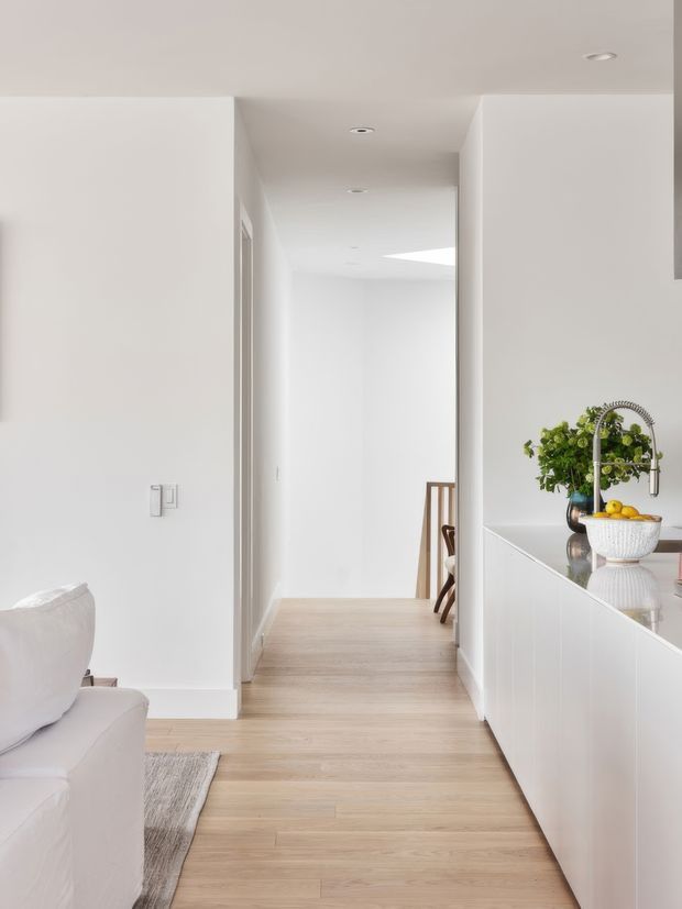 Bright minimalist interior view looking from a white living area toward a hallway and dining space, with a white kitchen island and integrated cabinetry on the right. Pale timber flooring runs throughout, and a skylight or clerestory opening illuminates the corridor. A partial white sofa and neutral rug are visible in the foreground, with a small plant and fruit bowl on the kitchen benchtop.