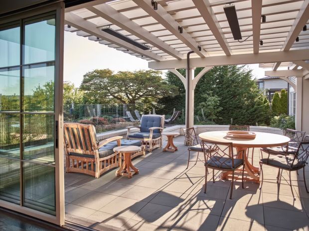 A spacious, sunlit outdoor terrace furnished with a round dining table and chairs alongside cushioned lounge seating. The terrace has large-format tiled paving, a glazed sliding door, and a substantial pergola with a translucent roof, exposed beams, integrated linear lighting, and ceiling fans. Established garden planting is visible beyond the glass balustrade.