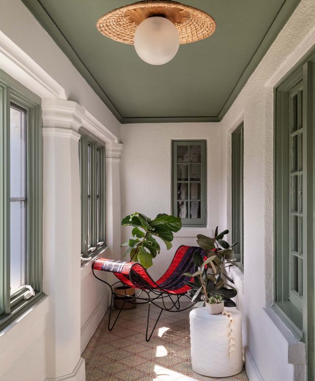 A narrow enclosed sunroom or covered verandah with white textured walls, sage-green ceiling and window/door frames, decorative white trim, and patterned tiled flooring. Tall windows line both sides, bringing in natural light. A colourful woven sling chair and potted plants create a relaxed indoor-outdoor seating area, with a woven ceiling pendant as the focal light.
