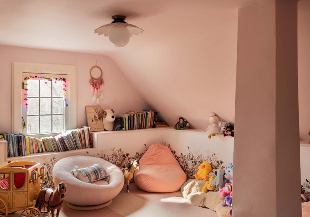 A cozy, playful children's room with soft pink walls and a sloped ceiling, filled with books, plush toys, colourful cushions, and low-level storage. A rounded lounge chair, floor beanbag, floral wall detail, decorative ceiling light, and a window with colourful trim create a warm, whimsical reading and play space.