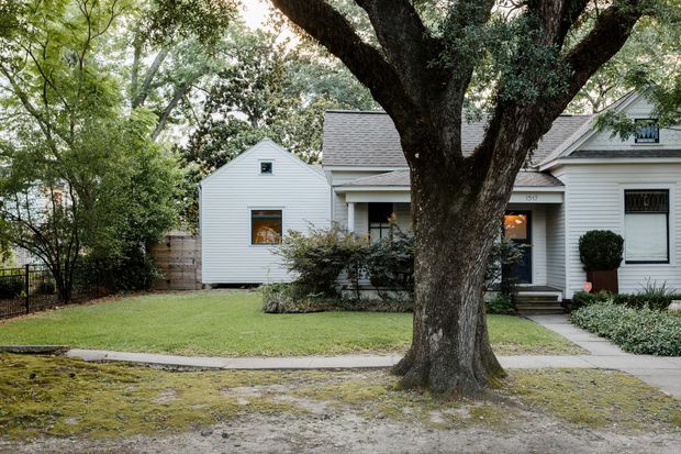 Front exterior view of a single-storey suburban house largely screened by a mature tree. The home features white horizontal weatherboard cladding, a grey shingle gabled roof, white-trimmed windows and a recessed entry porch. A small front lawn, concrete paths, garden beds and a low masonry boundary wall complete the setting.