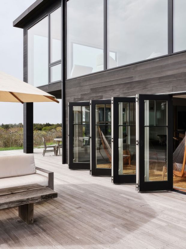 Exterior view of a contemporary two-storey home opening onto a broad timber deck. The facade combines dark horizontal timber cladding, large black-framed upper glazing, and a series of black-framed glazed folding or hinged doors. Outdoor lounge furniture and a cantilevered umbrella occupy the deck, with open landscape beyond.