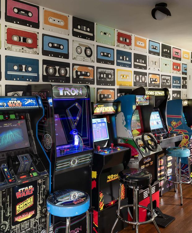 A vibrant retro arcade or games room filled with rows of classic arcade cabinets, including Star Wars, Tron, Defender and driving games. The walls are covered with colourful cassette-tape artwork, while stools, controllers and illuminated machine screens create a nostalgic, playful atmosphere. The room has timber flooring and a ceiling-mounted light fixture.