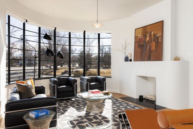 Bright, spacious modern living room with a curved wall of floor-to-ceiling black-framed glazing, a white sculptural fireplace, black leather seating, a patterned area rug, glass-and-metal coffee table, and a warm-toned abstract artwork above the mantel. The room has strong natural light and views of trees and a residential street outside.