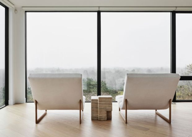 A minimalist living area with two upholstered lounge chairs positioned in front of expansive black-framed floor-to-ceiling windows. A small textured wood side table sits between the chairs, while pale herringbone timber flooring and white walls create a bright, serene setting. A hazy landscaped view is visible beyond the glazing.