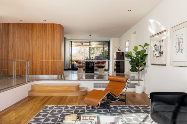 Bright, open-plan modern living space with a warm timber-paneled curved partition and matching timber flooring. The room includes a sunken seating area, a leather lounge chair, patterned rug, glass balustrade, framed artwork, indoor plant, and a dining or kitchen zone visible toward the rear through an open connection. Large glazing brings in natural light and views of trees.