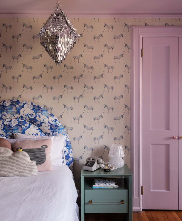 A charming, eclectic bedroom corner with a single bed partially visible at left, layered pillows, a blue-and-white floral headboard or bedspread, patterned animal wallpaper, and a small teal bedside cabinet. A sparkling metallic pendant light hangs from the ceiling, while the door and trim are painted soft pink. Timber flooring is visible at the bottom.