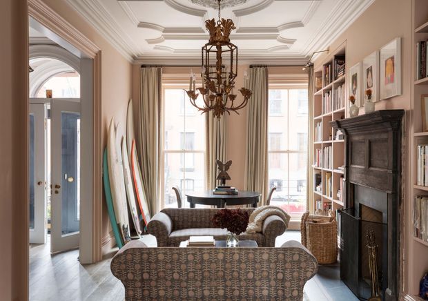 Elegant, traditionally detailed living room viewed from behind a patterned sofa, featuring a carved dark fireplace, full-height built-in bookshelves, ornate chandelier, decorative ceiling mouldings, tall curtained windows, and a small seating area. A blue-grey entry door and surfboards are visible through the opening to the left.