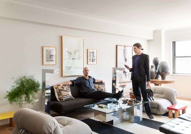 Bright, refined contemporary living room with pale walls, decorative crown moulding, large windows, layered abstract artwork, a dark sofa, sculptural lounge chairs, and a transparent glass coffee table. Two people are present, and the room includes a console with decorative vessels and a patterned area rug.