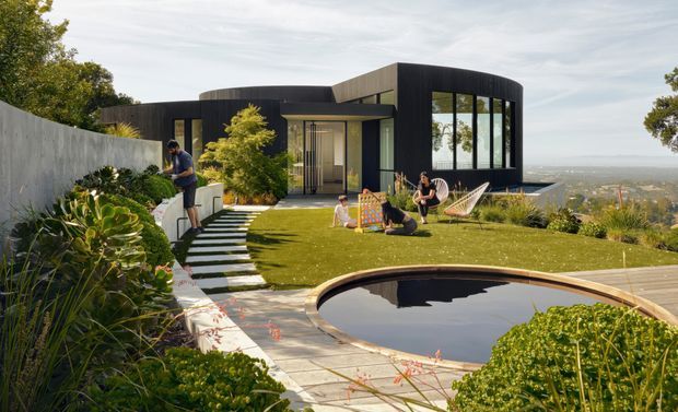 A contemporary dark-clad curved-form house set on a landscaped sloping site, viewed from the garden. The property features extensive glazing, a manicured lawn, stepping-stone path, planted retaining edges, timber decking and a reflective swimming pool in the foreground, with a distant coastal or urban outlook beyond.